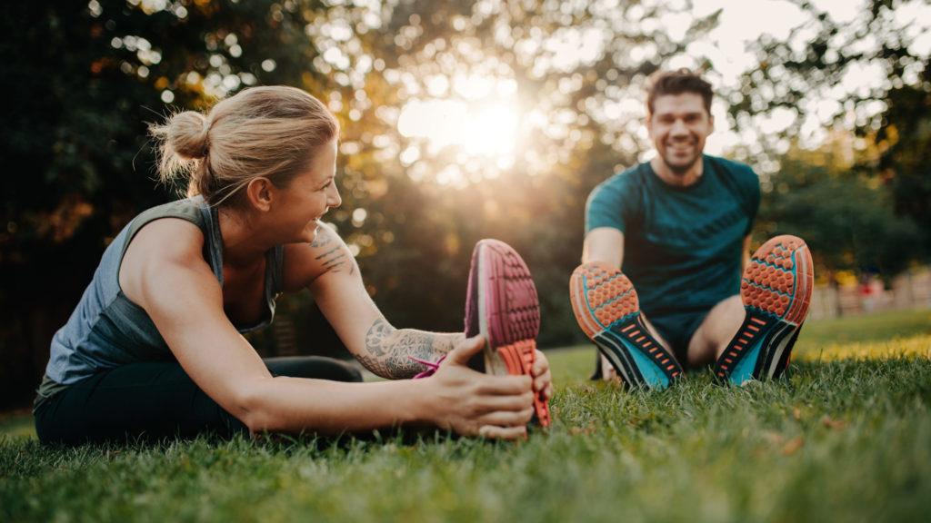 male and female enjoying exercise together outdoors by stretching on the grass on a sunny day