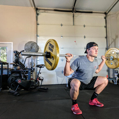 A man is doing a barbell squat in a garage gym. He is wearing a gray T-shirt, black shorts, red shoes, and a black cap worn backward. Exercise equipment, like a stationary bike and weights, is visible in the background.